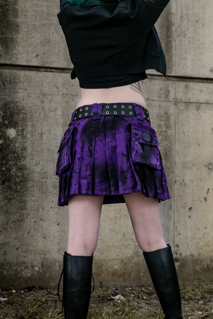 Back view of a person wearing a purple and black pleated mini kilt with black belt and boots against a concrete wall.