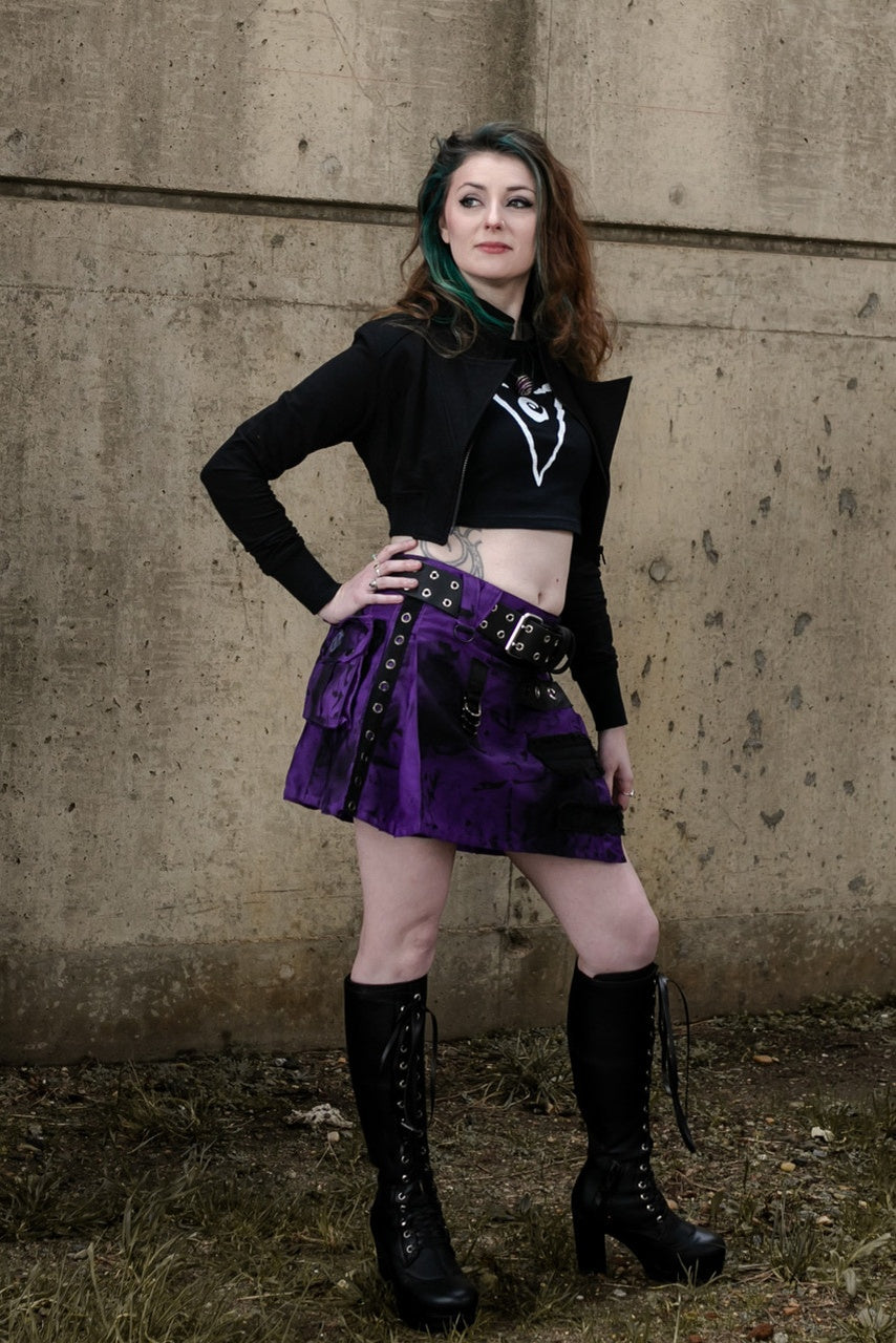 Woman in a black top and purple and black pleated mini kilt standing against a concrete wall.