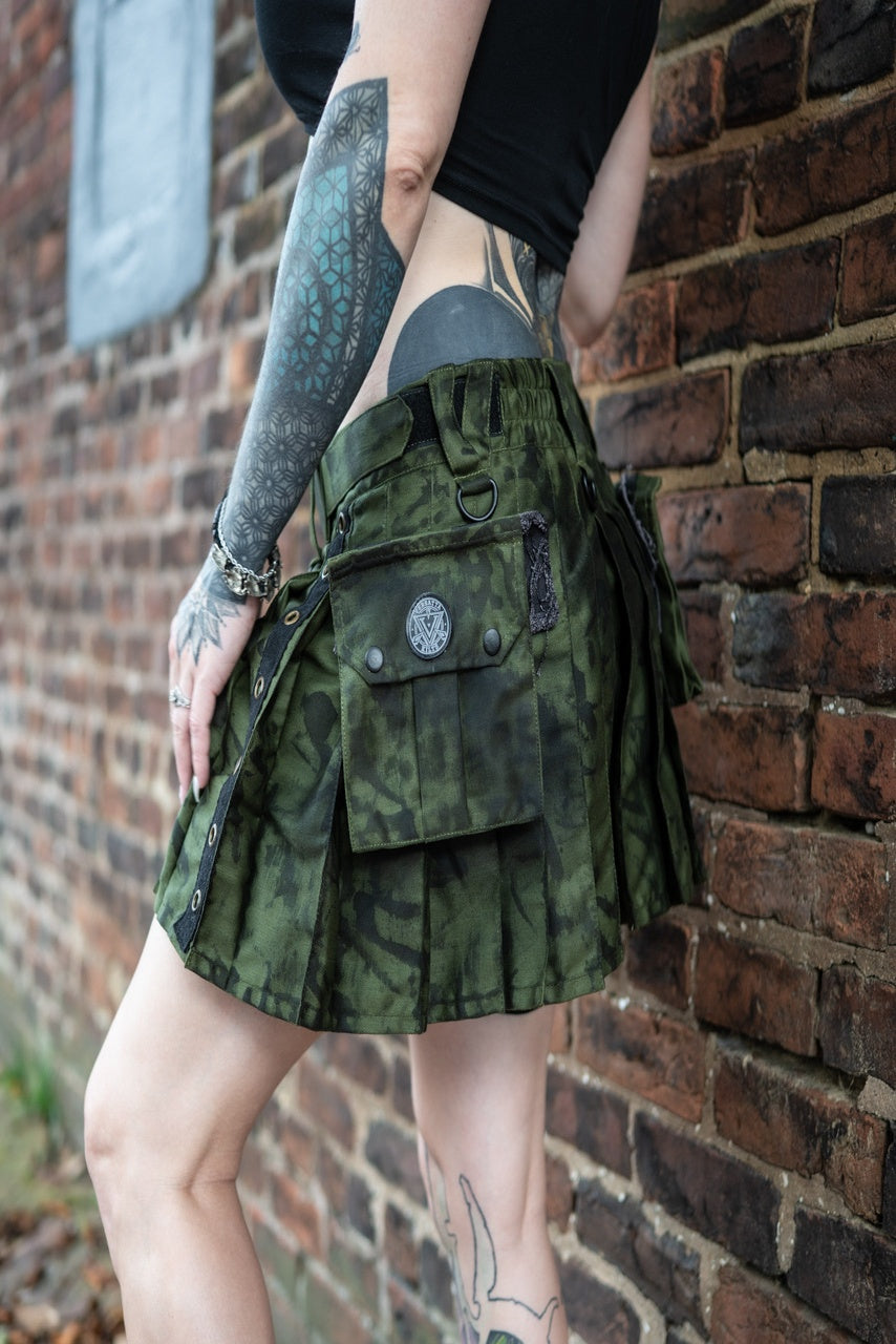 Person wearing a dark green pleated kilt with black patterns against a brick wall.