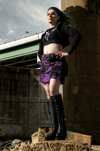 Person wearing a black top, purple and black mini kilt, and black boots standing on a concrete block with a bridge in the background.